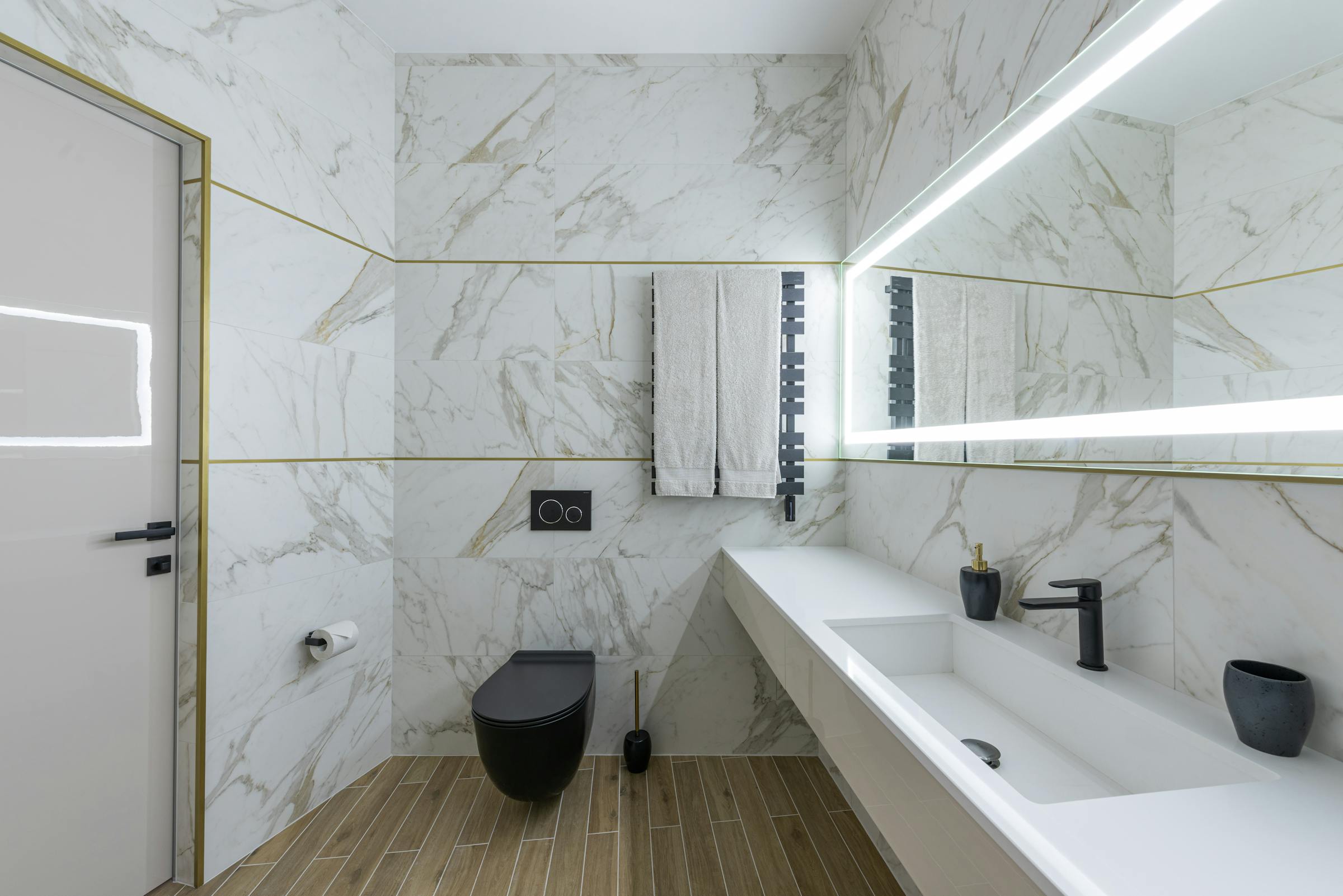Marble-clad bathroom with warm wood floor, brass fittings, and a linear LED mirror